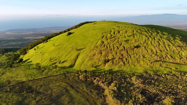Aerial view of Puu Waawaa cinder cone featuring lush green slopes and volcanic ridges overlooking lava fields and the distant ocean in Hawaii County, Hawaii, United States.