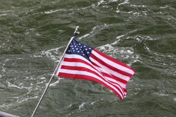 Fototapeta premium United States flag blowing in the wind on a metal pole above the rippled surface of the Gastineau Channel in Alaska