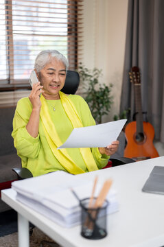 Happy elderly senior old asian woman holding document talking on phone call at table in home office
