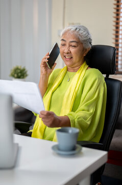 Smiling elderly senior old asian woman holding papers talking on phone call at table in home office
