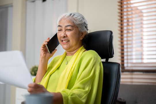 Happy elderly senior old asian woman holding documents talking on phone call at table in home office