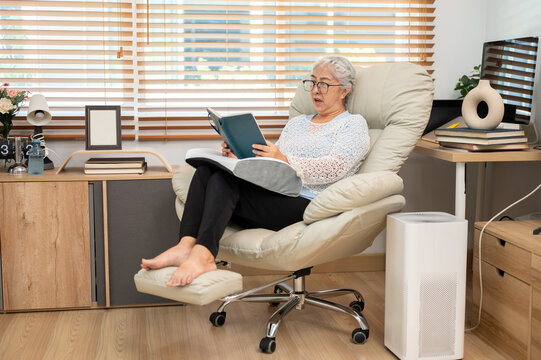 Retired elderly old asian woman reading a book mouth gaping on sofa armchair in living room at home.