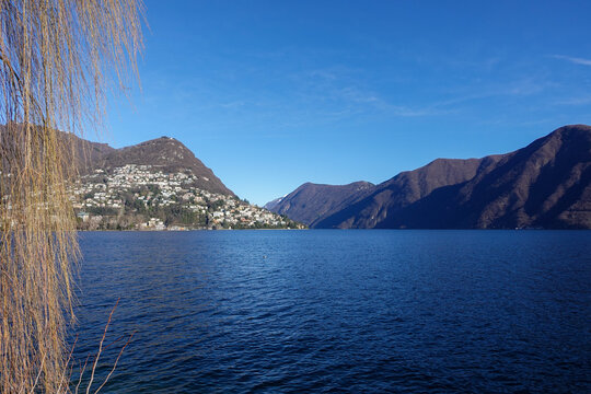 Under clear daylight, Lake Lugano in Southern Switzerland, shimmers between mountain slopes, reflecting quiet villages, deep blue water, and the calm rhythm of the alpine landscape.