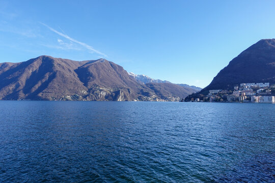 Under clear daylight, Lake Lugano in Southern Switzerland, shimmers between mountain slopes, reflecting quiet villages, deep blue water, and the calm rhythm of the alpine landscape.