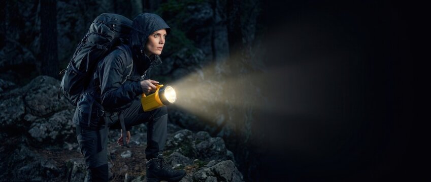 Woman explorer navigating dark wilderness at night, searching with a powerful flashlight