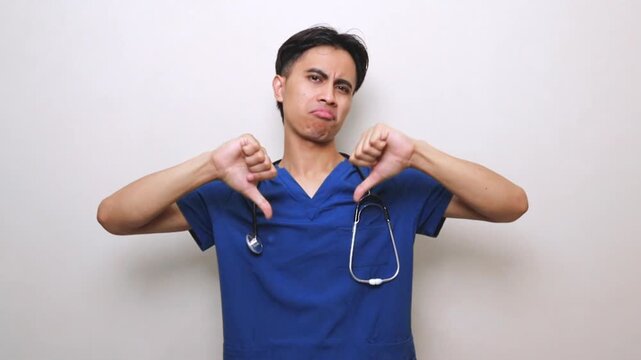Asian male nurse in blue medical scrubs showing thumb down gesture as sign of disapproval or medical warning