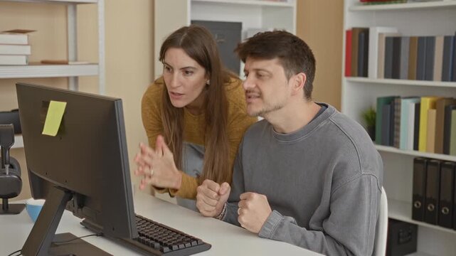 Man at computer with hands pressed together in praying gesture and woman leaning with hands near mouth, office desk and bookshelf visible; concern teamwork problem solving.