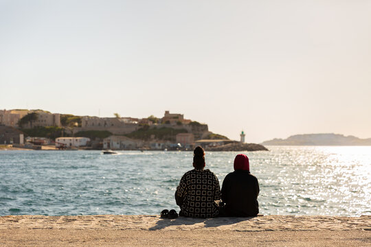 Deux femmes vues de dos assises sur une digue face &agrave; la mer et d'un petit port en face au loin, une femme porte un voile sur la t&ecirc;te, lumi&egrave;re douce de fin d'apr&egrave;s-midi, ville de Marseille
