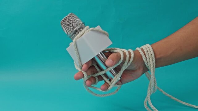 Hand Struggles To Hold News Microphone Tightly Bound With Rope On Blue Background Representing Suffocation Of Free Speech And Press