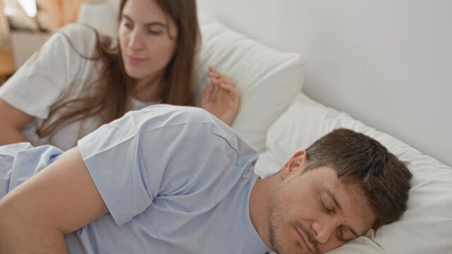 Man lies on bed pillow looking up as woman leans over smiling and touches his shoulder and face in a bedroom inside a building; affection warmth.