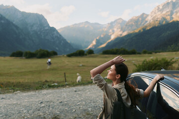 Naklejka premium Woman beside a car in a mountain meadow, enjoying fresh air and wide landscape, conveys travel freedom and outdoor lifestyle in a sunny rural scene