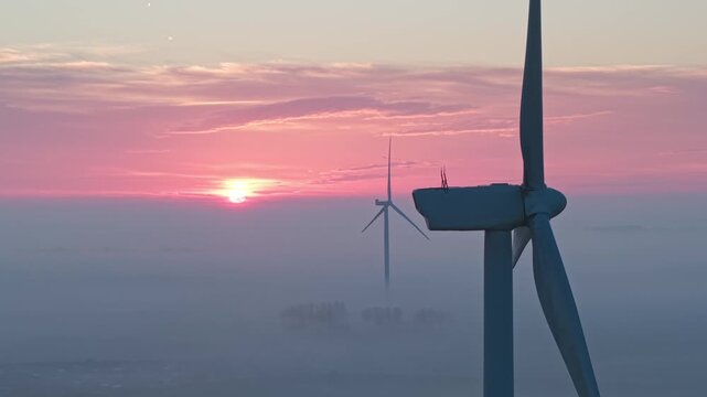 Aerial view of a single wind turbine emerging from thick morning fog under a pink sunset sky in Vrouwenpolder, Zeeland, Netherlands.