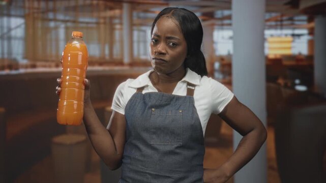 Woman wearing denim apron holds large orange juice bottle in hand in building cafe interior, examining cap and label with raised eyebrow; disapproval concern.