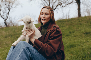 Naklejka premium Cheerful woman outdoors, seated on grass, holding a young goat kid, warm jacket, gentle breeze, sunlit scene, natural colors, field background, candid lifestyle moment, calm connection with animals