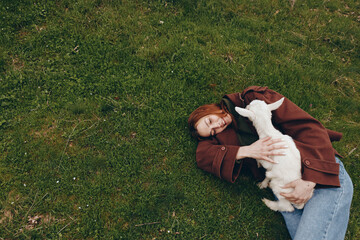 Naklejka premium Young woman with red hair lies on green grass holding a white baby goat and smiling happily in a relaxed outdoor lifestyle scene.