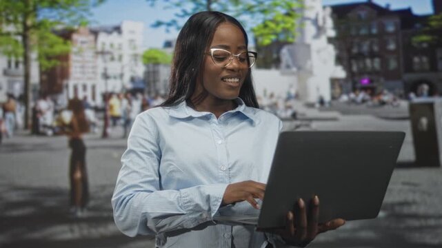 Woman holding laptop and typing on street amid a busy city square with blurred pedestrians and buildings; confidence.