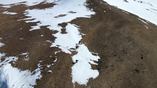 Aerial view of hikers trekking along the snowy Moskardshnjukar Trail ridge with brown rocky terrain and snow patches in Mosfellsdalur, Reykjavikurborg, Iceland.