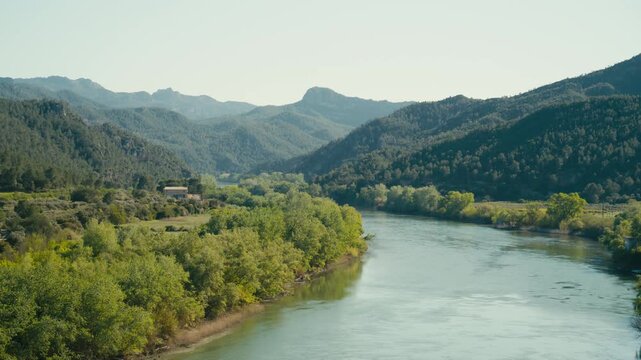 The Ebro River winds through green riverbanks and forested hills in Spain, creating a broad natural landscape with calm water and distant mountain ridges.