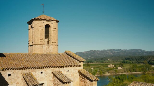 A historic bell tower rises above Miravet, Spain, with tiled rooftops, the Ebro valley, and distant mountains creating a warm and scenic medieval setting.