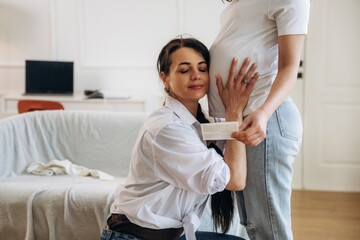 Pregnant woman in casual attire stands while another woman kneels beside her, gently touching her belly in a bright, modern living room setting © standret