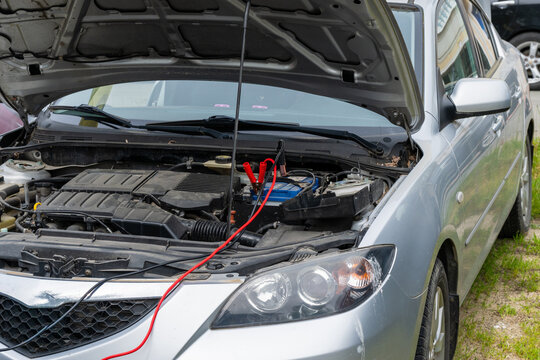 A silver car parked outdoors with its hood propped open and jumper cables attached to its battery in preparation for a jump-start.
