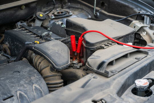 A bright red jumper cable clamp is securely attached to a battery terminal within a dusty car engine bay, preparing for a jump-start.