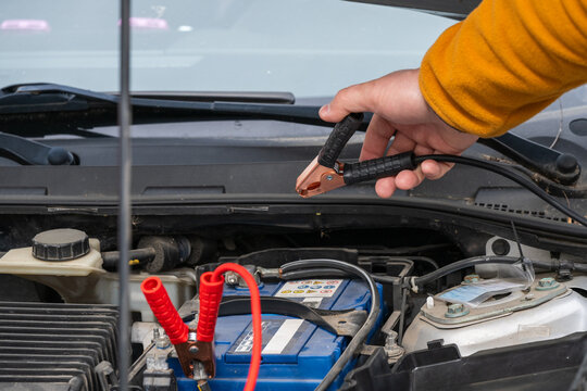 a person's hand holding a black jumper cable clamp over a car engine bay, preparing to connect it alongside an already attached red clamp to jump-start a dead battery.