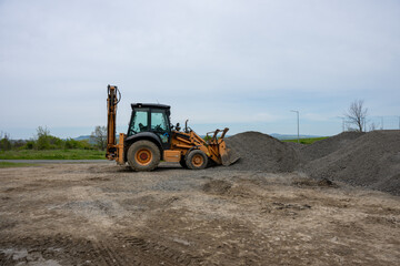 Obraz premium A yellow backhoe loader uses its front bucket to push a large pile of gravel on a dirt lot under a cloudy sky.
