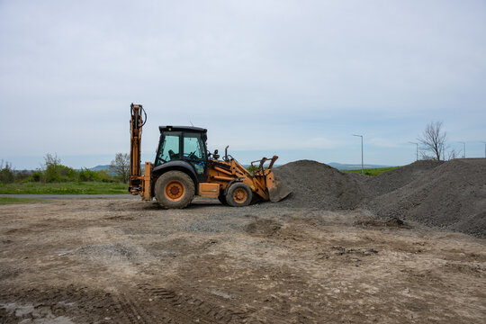 A yellow backhoe loader uses its front bucket to push a large pile of gravel on a dirt lot under a cloudy sky.