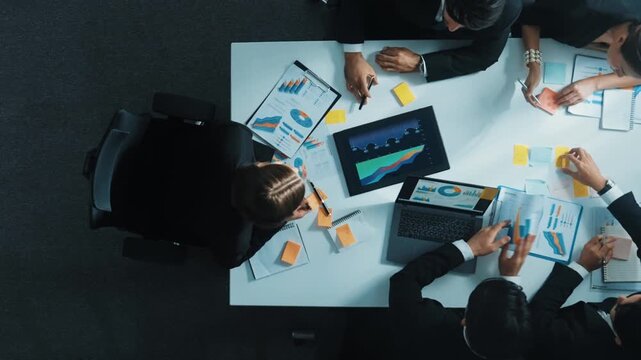 Top down aerial view of diverse business people discuss and brainstorm marketing idea while looking at financial graph at table with marketing document and sticky notes scatter around. Directorate.