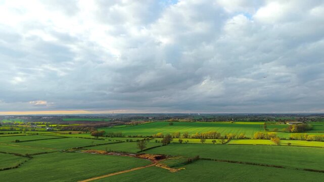 Bright agricultural fields forming geometric patterns across rolling countryside under textured cloudy sky. Natural light illuminating sections of farmland and tree lines. Peaceful rural environment
