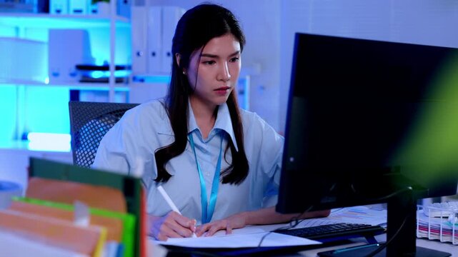 Office woman employee at computer desk handling paperwork and corporate document work, focused business worker typing and writing notes during night overtime shift in modern workplace with blue light