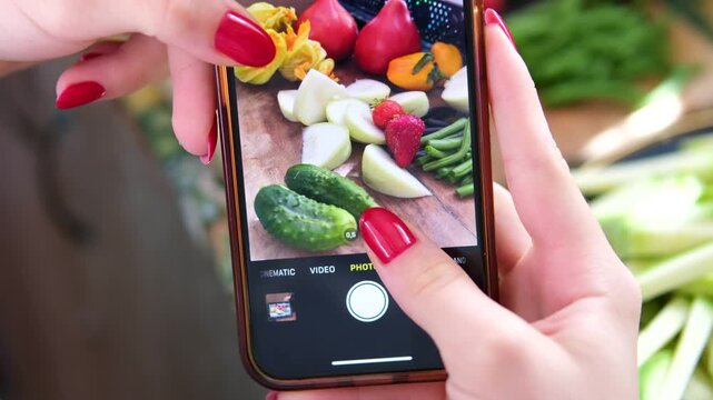 Female hands with red manicure record a wooden crate filled with fresh vegetables using a smartphone. Cucumbers, zucchini with yellow blossoms, pink tomatoes, onions. 
