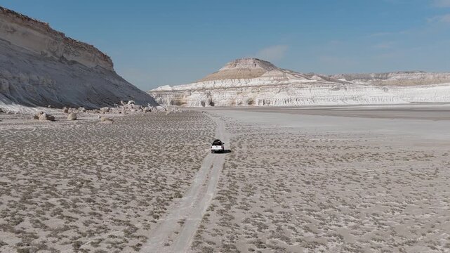 Aerial view of an off-road vehicle driving on a dirt track through the white limestone cliffs and salt flats of Tuzbair in Mangystau District, Mangystau Region, Kazakhstan.