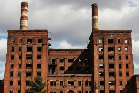 Old brick factory buildings with broken windows, tall smokestacks stand against cloudy sky. Abandoned industrial complex shows signs of decay, neglect. Nature begins reclaiming unused structures.