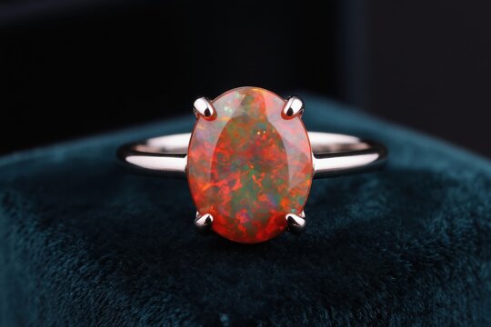 Oval cut fire opal ring on silver band. Gemstone glows with orange red fire and green flashes. Minimalist jewelry on dark velvet, luxury accessory detail. Elegant fashion engagement.