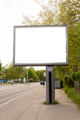 Obraz premium A large empty billboard is positioned next to a road. The surrounding area features green trees and minimal traffic. It is a clear day with sunlight