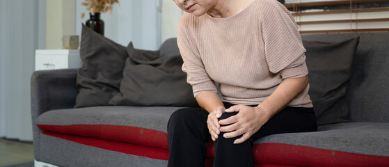 Fototapeta premium Elderly woman suffering from knee pain osteoarthritis. Close up senior woman holding her knee due to physical injury while sitting alone at home. rheumatoid arthritis disease in Adult