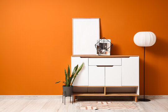 Interior of living room with lamp, houseplant and home decor on white commode near orange wall