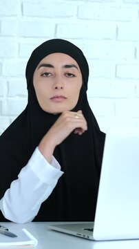 Young islamic woman wearing a black hijab sitting at her desk in the office, thoughtfully looking at the camera while working on her laptop computer with a focused expression