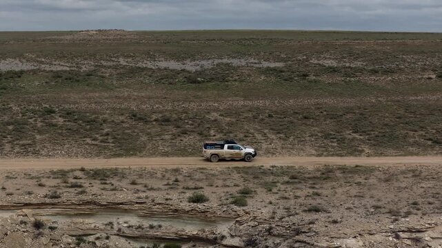 Aerial view of an off-road vehicle driving on a dirt road through the rocky desert landscape of Bozjyra with a cloudy sky in Aktau, Mangystau Region, Kazakhstan.