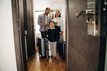Family of three with luggage stands at hotel room entrance, smiling and ready for their stay, showcasing modern interior design and wooden flooring © standret