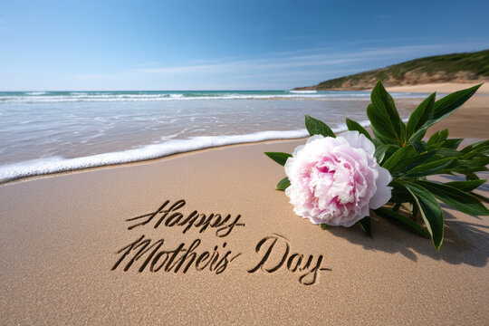 Happy Mothers Day written on sandy beach with pink peony flower, ocean wave, blue sky, summer, peaceful, celebration, nature