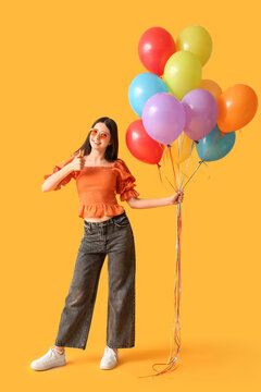 Beautiful teenage girl in sunglasses with colorful air balloons showing thumb-up gesture on yellow background