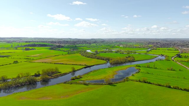 Flowing river curves through vibrant agricultural land with hedgerows and scattered trees shaping terrain. High altitude drone view farmland textures and waterway patterns across open countryside