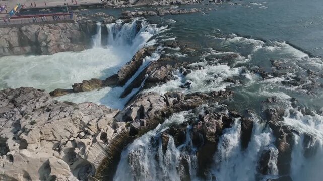 Dhuandhar Falls rushes over dark volcanic rock formations creating multiple cascades into turquoise pools. Aerial shot reveals the dramatic gorge terrain and powerful water flow of Narmada River.