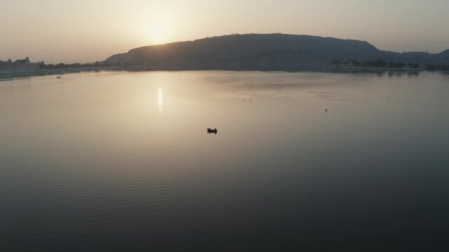 Fishing boat drifts across the calm waters of Lake Pichola during golden hour with silhouetted hills in the background. Slow lateral pan left reveals lakeside architecture and captures Udaipur city.