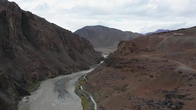 Ladakh's Indus River winds through a high-altitude desert canyon flanked by barren rocky slopes. Smooth drone movement reveals the turquoise glacial river and remote Himalayan snow capped mountains.