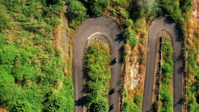 Motorcycles traverse winding hairpin curves on a mountain road in Yercaud, Tamil Nadu. Aerial view captures the serpentine asphalt path cutting through dense green forest vegetation, Salem.