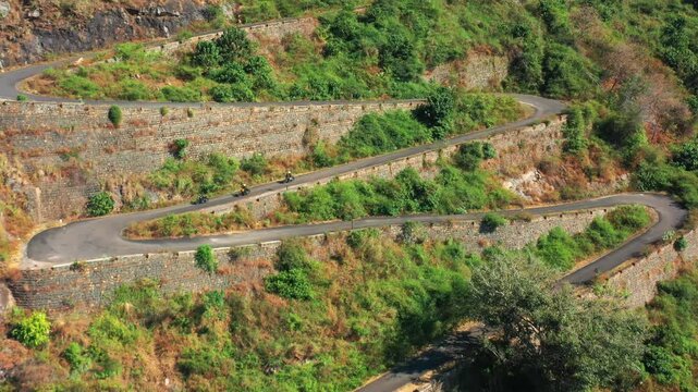 Yercaud Ghat Road winds through hillside terrain with multiple hairpin turns and stone retaining walls. Aerial view captures motorcycles descending the serpentine road with sparse vegetation.